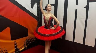 Young ballerina in a red and black tutu strikes a pose, holding a tambourine overhead backstage.