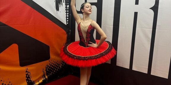 Young ballerina in a red and black tutu strikes a pose, holding a tambourine overhead backstage.