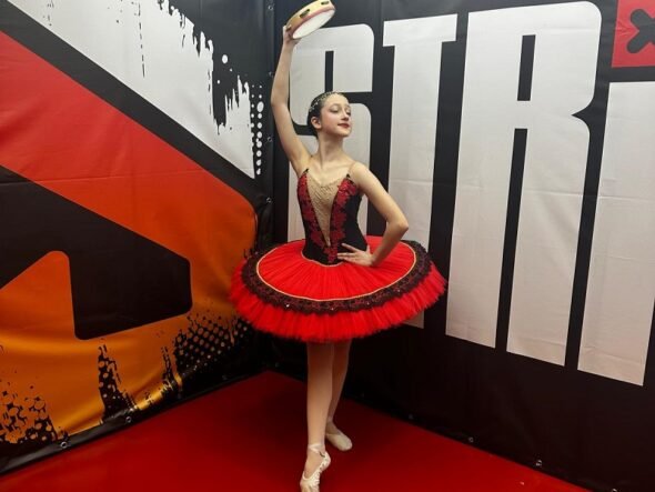 Young ballerina in a red and black tutu strikes a pose, holding a tambourine overhead backstage.