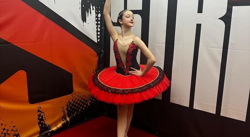 Young ballerina in a red and black tutu strikes a pose, holding a tambourine overhead backstage.