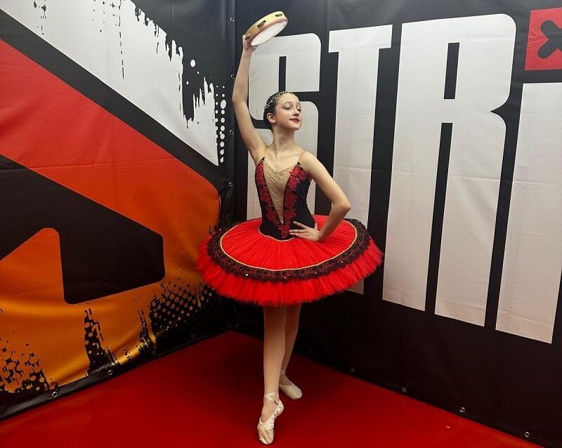 Young ballerina in a red and black tutu strikes a pose, holding a tambourine overhead backstage.