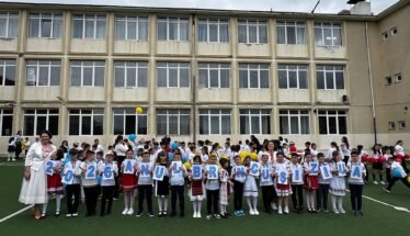 Group of primary school children in white shirts and traditional outfits, standing on a field in front of a beige school building, each holding a card with a letter to form a message.