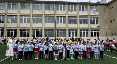 Group of primary school children in white shirts and traditional outfits, standing on a field in front of a beige school building, each holding a card with a letter to form a message.