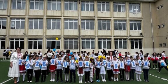 Group of primary school children in white shirts and traditional outfits, standing on a field in front of a beige school building, each holding a card with a letter to form a message.