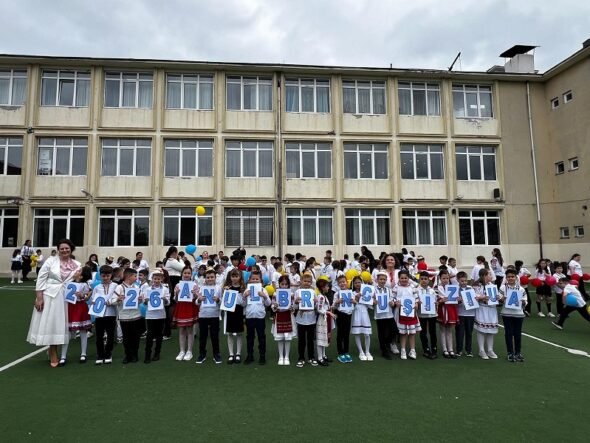 Group of primary school children in white shirts and traditional outfits, standing on a field in front of a beige school building, each holding a card with a letter to form a message.