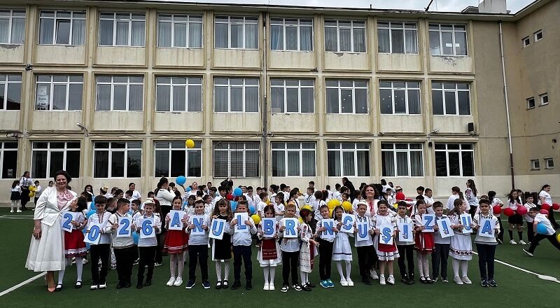 Group of primary school children in white shirts and traditional outfits, standing on a field in front of a beige school building, each holding a card with a letter to form a message.
