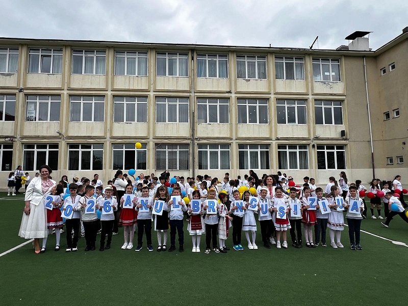 Group of primary school children in white shirts and traditional outfits, standing on a field in front of a beige school building, each holding a card with a letter to form a message.