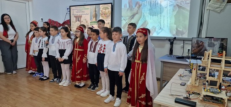 Group of children in traditional embroidered outfits standing in a classroom, ready to perform for an audience near a projector screen and tables with equipment.