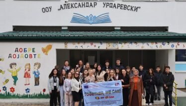 Group of students and teachers posing outside a school, holding a banner that reads 'TOGETHER, WE CAN!', with colorful wall art and the school's façade in the background.