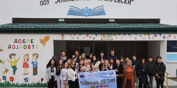 Group of students and teachers posing outside a school, holding a banner that reads 'TOGETHER, WE CAN!', with colorful wall art and the school's façade in the background.