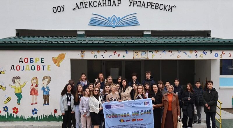 Group of students and teachers posing outside a school, holding a banner that reads 'TOGETHER, WE CAN!', with colorful wall art and the school's façade in the background.
