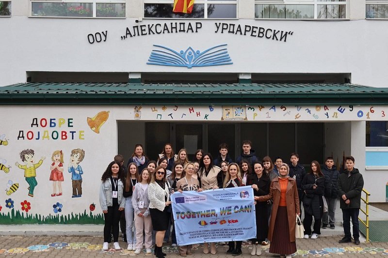 Group of students and teachers posing outside a school, holding a banner that reads 'TOGETHER, WE CAN!', with colorful wall art and the school's façade in the background.