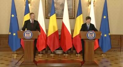 Two men in suits speak at podiums in a formal government room, with EU and Romanian flags behind them.