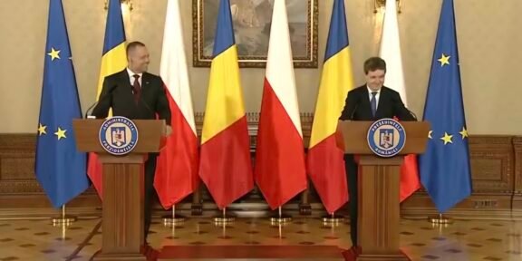 Two men in suits speak at podiums in a formal government room, with EU and Romanian flags behind them.