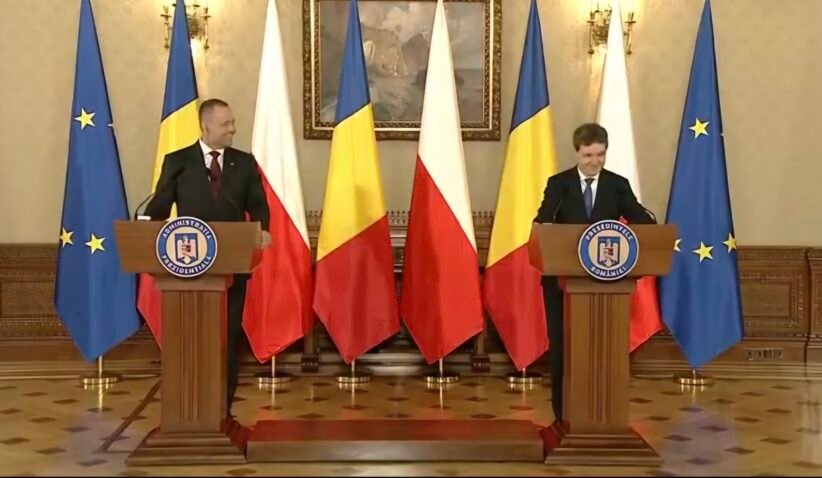 Two men in suits speak at podiums in a formal government room, with EU and Romanian flags behind them.