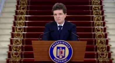 Man in a dark suit delivering a formal speech at a wooden podium with a round blue-and-white seal, in front of a red-carpeted staircase backdrop.