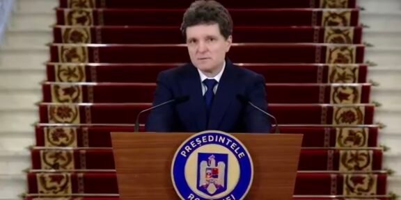 Man in a dark suit delivering a formal speech at a wooden podium with a round blue-and-white seal, in front of a red-carpeted staircase backdrop.