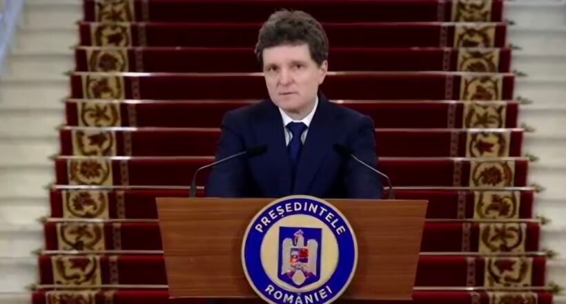 Man in a dark suit delivering a formal speech at a wooden podium with a round blue-and-white seal, in front of a red-carpeted staircase backdrop.