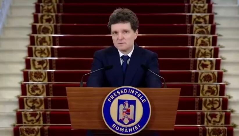 Man in a dark suit delivering a formal speech at a wooden podium with a round blue-and-white seal, in front of a red-carpeted staircase backdrop.