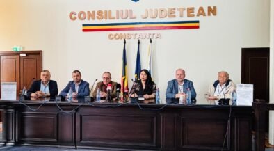 Panel of seven officials seated behind a long dark wood desk in a government chamber, with Romanian flags and 'Consiliul Judetean Constanta' on the wall.