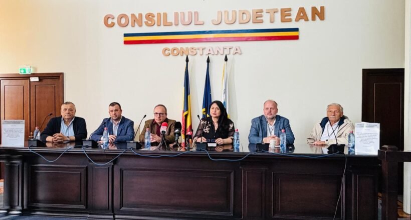 Panel of seven officials seated behind a long dark wood desk in a government chamber, with Romanian flags and 'Consiliul Judetean Constanta' on the wall.