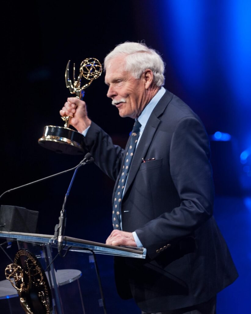 Older man in a dark suit at a podium holding an Emmy Award, blue stage lighting in the background
