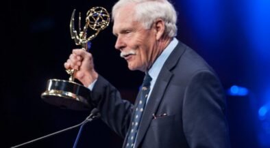 Older man in a dark suit at a podium holding an Emmy Award, blue stage lighting in the background