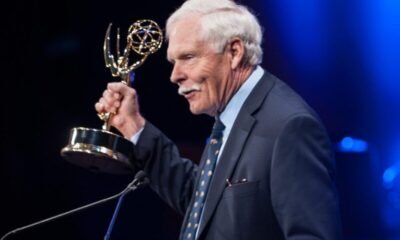 Older man in a dark suit at a podium holding an Emmy Award, blue stage lighting in the background