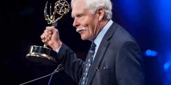 Older man in a dark suit at a podium holding an Emmy Award, blue stage lighting in the background