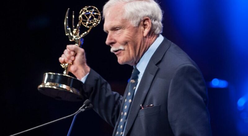 Older man in a dark suit at a podium holding an Emmy Award, blue stage lighting in the background