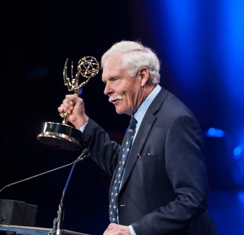 Older man in a dark suit at a podium holding an Emmy Award, blue stage lighting in the background