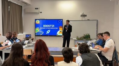 Presenter in a dark suit speaks at the front of a classroom-style room; BOOST21 slide on a large screen behind him while attendees with laptops listen around tables.