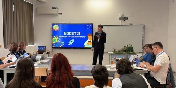 Presenter in a dark suit speaks at the front of a classroom-style room; BOOST21 slide on a large screen behind him while attendees with laptops listen around tables.