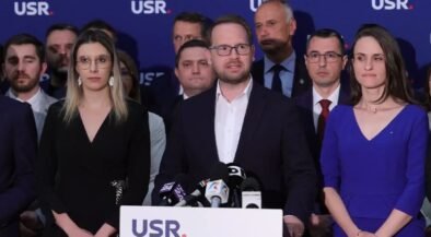 Man in a dark suit speaks at a podium with microphones, flanked by a woman in black on his left and a woman in blue on his right, supporters in the background with a blue USR backdrop.