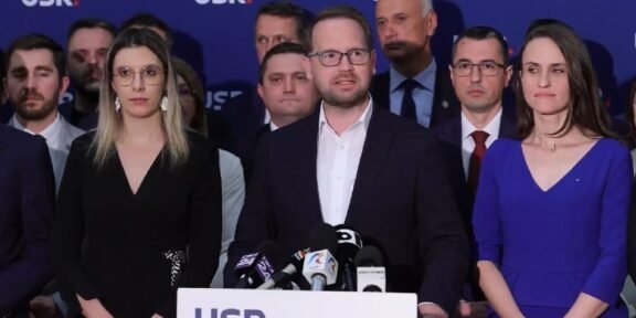 Man in a dark suit speaks at a podium with microphones, flanked by a woman in black on his left and a woman in blue on his right, supporters in the background with a blue USR backdrop.