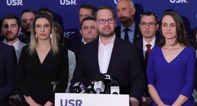 Man in a dark suit speaks at a podium with microphones, flanked by a woman in black on his left and a woman in blue on his right, supporters in the background with a blue USR backdrop.