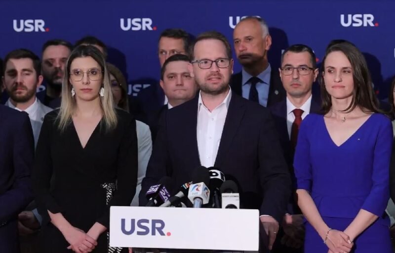Man in a dark suit speaks at a podium with microphones, flanked by a woman in black on his left and a woman in blue on his right, supporters in the background with a blue USR backdrop.