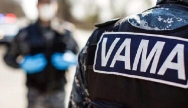 Close-up of a tactical uniform with a VAMA patch; blurred officer with blue gloves and police lights in the background.
