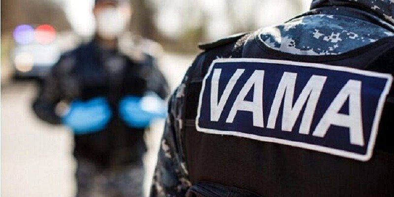 Close-up of a tactical uniform with a VAMA patch; blurred officer with blue gloves and police lights in the background.