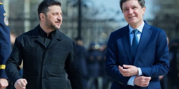 Two men walk side by side outdoors, one in a dark jacket and the other in a blue suit, smiling as they pass a crowd of onlookers.