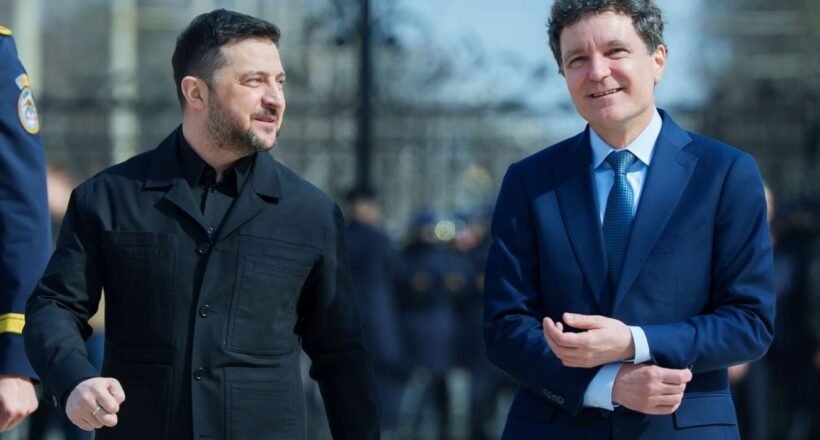 Two men walk side by side outdoors, one in a dark jacket and the other in a blue suit, smiling as they pass a crowd of onlookers.