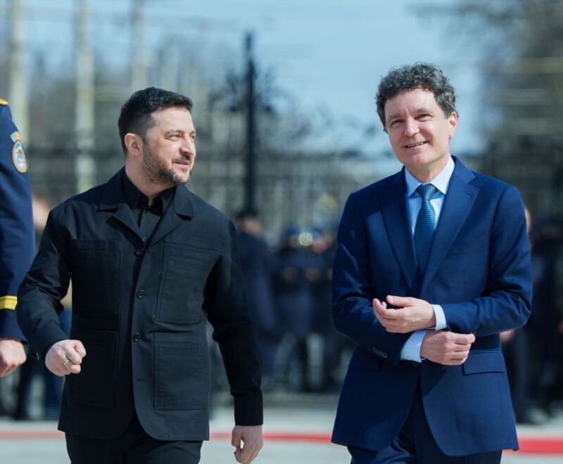 Two men walk side by side outdoors, one in a dark jacket and the other in a blue suit, smiling as they pass a crowd of onlookers.
