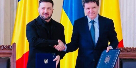 Two men in formal attire shake hands over a table, each holding a blue folder with a crest, Ukrainian flags in the background.