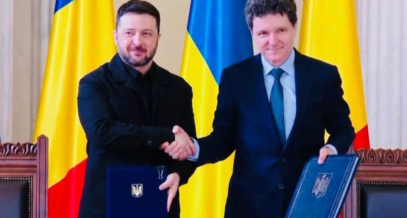 Two men in formal attire shake hands over a table, each holding a blue folder with a crest, Ukrainian flags in the background.