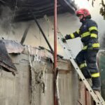 Firefighter in turnout gear on a ladder beside a smoke-filled, fire-damaged building exterior.