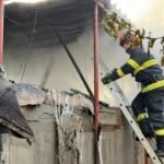 Firefighter in turnout gear on a ladder examining a burnt building with smoke and charred debris.