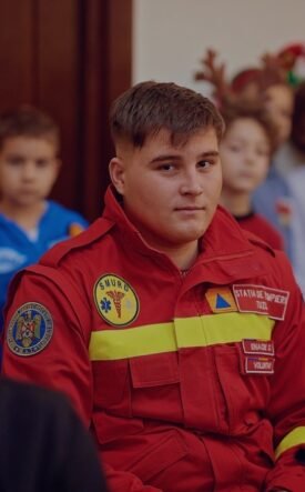 Young man in a red emergency responder uniform sits among a group at an indoor event, patches visible on the chest.