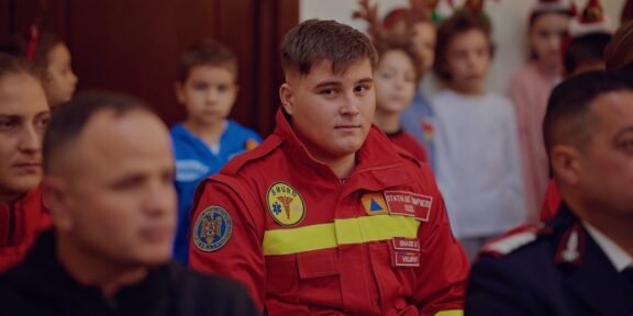 Young man in a red emergency responder uniform sits among a group at an indoor event, patches visible on the chest.