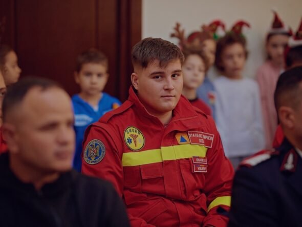 Young man in a red emergency responder uniform sits among a group at an indoor event, patches visible on the chest.
