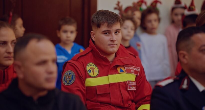 Young man in a red emergency responder uniform sits among a group at an indoor event, patches visible on the chest.